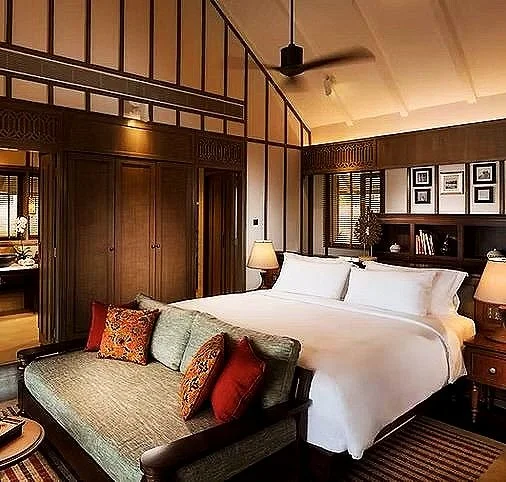 Hotel bedroom with a large bed, decorative pillows, bedside lamps, and a sitting area with a sofa, wood-paneled walls, and framed photos.