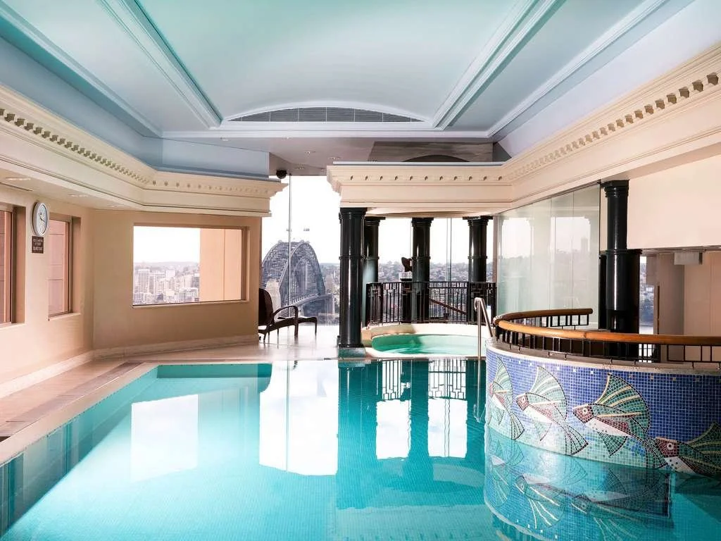 Aerial view of a tropical beach resort with a sandy shoreline, lounge chairs, lush green hills, and multiple buildings with red roofs near the coast.
