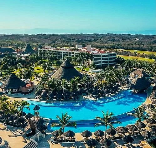 Resort with a large curved swimming pool, surrounded by palm trees, thatched-roof huts, and a hotel building in the background, set in a tropical landscape.