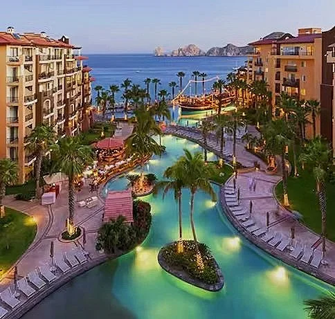 Aerial view of a luxury waterfront resort with a swimming pool, palm trees, multi-story buildings, and ocean in the background