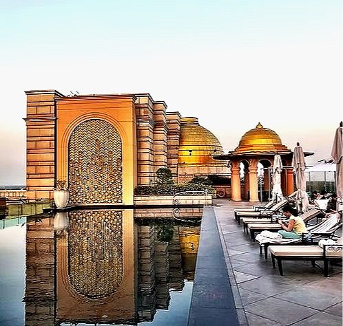 Luxury rooftop pool area with a reflection of ornate building facades, domed structures, and lounge chairs with a person sitting, during sunset.
