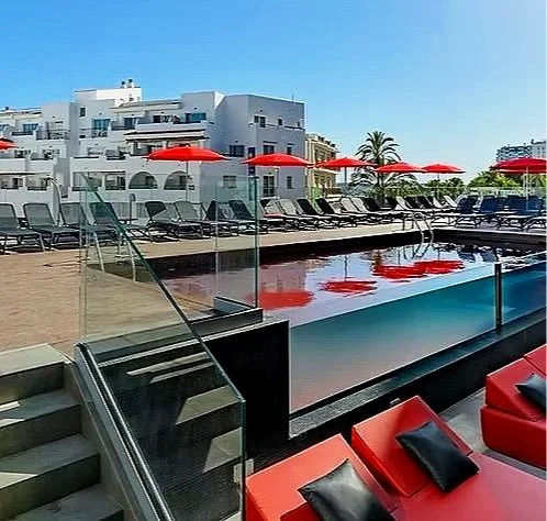 Empty swimming pool area with red umbrellas, lounge chairs, and modern white buildings in the background.