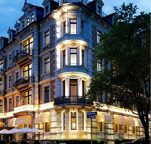 Elegant multi-story historic building with detailed architecture, illuminated at night, with balconies and a corner turret, surrounded by trees.
