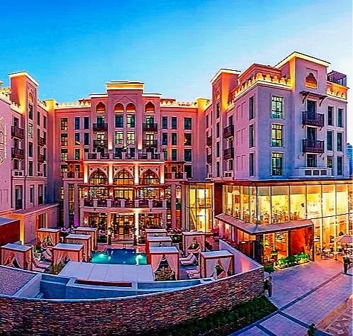 A large, elegant hotel or apartment complex with pink and beige exterior, multiple balconies, and a central courtyard featuring a swimming pool and outdoor seating areas, illuminated at dusk.