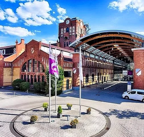 A historic red-brick building with a tall clock tower and a modern curved glass entrance, situated in a paved courtyard with flags on poles and parked cars.
