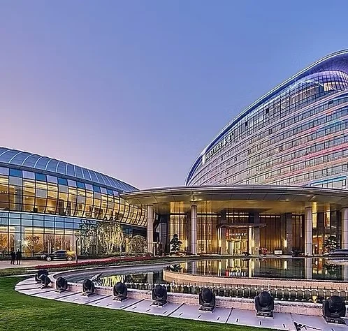 Modern architectural building with curved glass façade, large circular entrance, and a reflecting pool with stage lights in front.