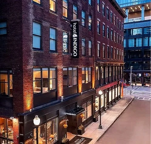 Exterior view of a brick hotel called Hotel Indigo in an urban area at dusk. The building has lit windows and street lamps, with a sidewalk and crosswalk visible.