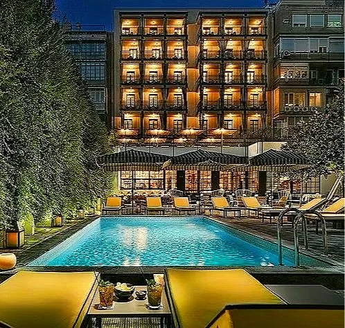 Nighttime view of a lit hotel with a pool in front, lounge chairs, and tables with drinks, surrounded by greenery.