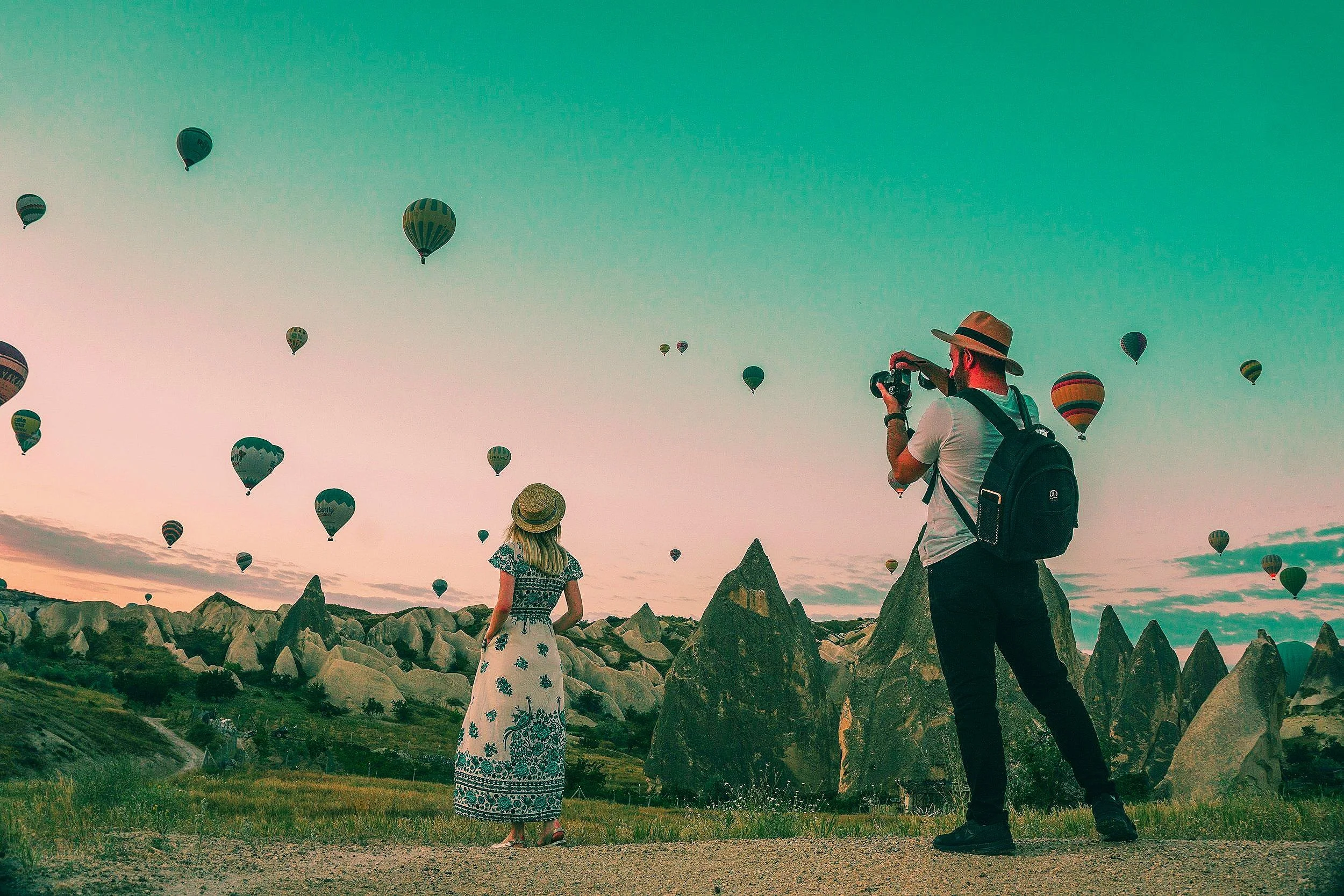 A woman and a man are standing outdoors during sunset, watching hot air balloons in the sky with rocky formations in the background. The man is taking a photo with a camera.