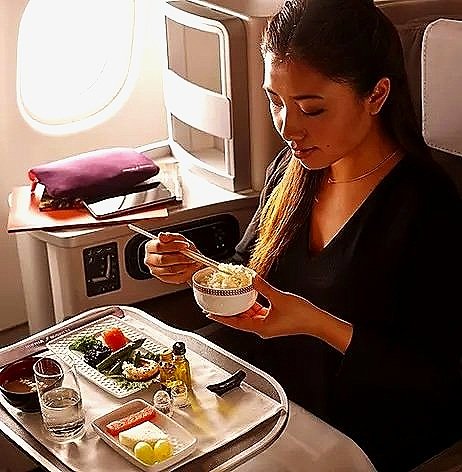 A woman sitting in an airplane seat eating a bowl of food with chopsticks, with a tray table holding a glass of water, sushi, and condiments.