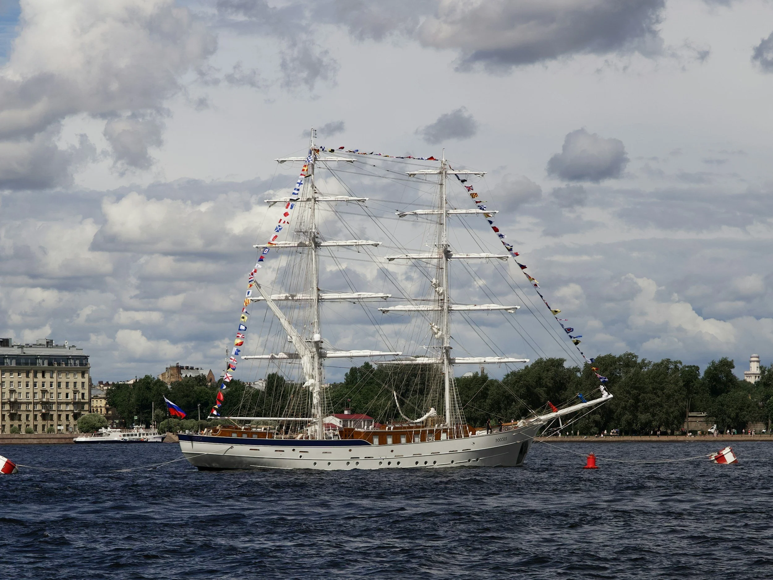 A large white sailing ship decorated with colorful flags is on a river, with an urban cityscape and trees in the background under a cloudy sky.
