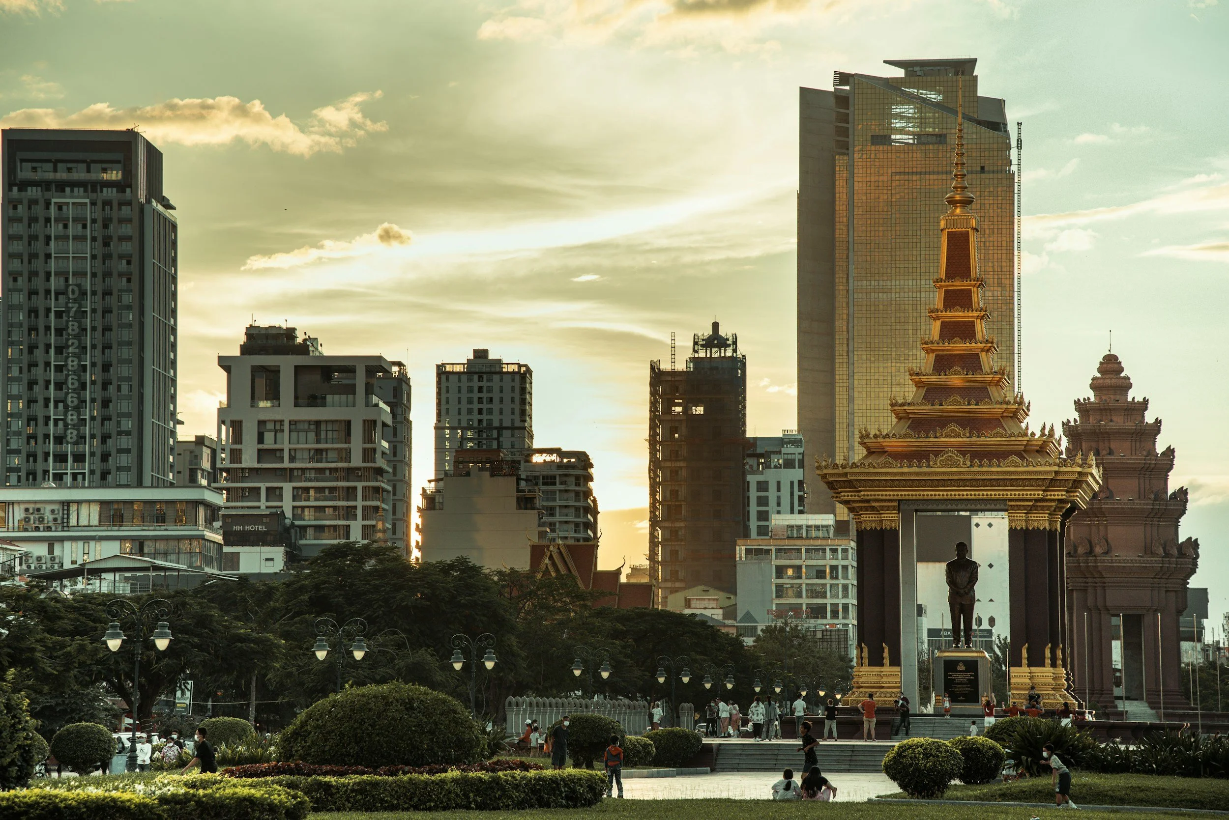 Cityscape of a downtown area with tall modern buildings, an historic church with a green dome, and a park with colorful trees in the foreground at dusk