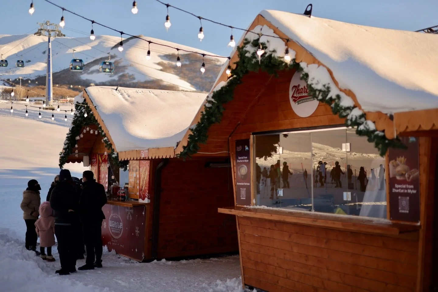A winter scene at a ski resort with wooden food booths decorated with holiday garlands, snow-covered roofs, and string lights, with people in winter clothing standing nearby and ski lifts in the background.