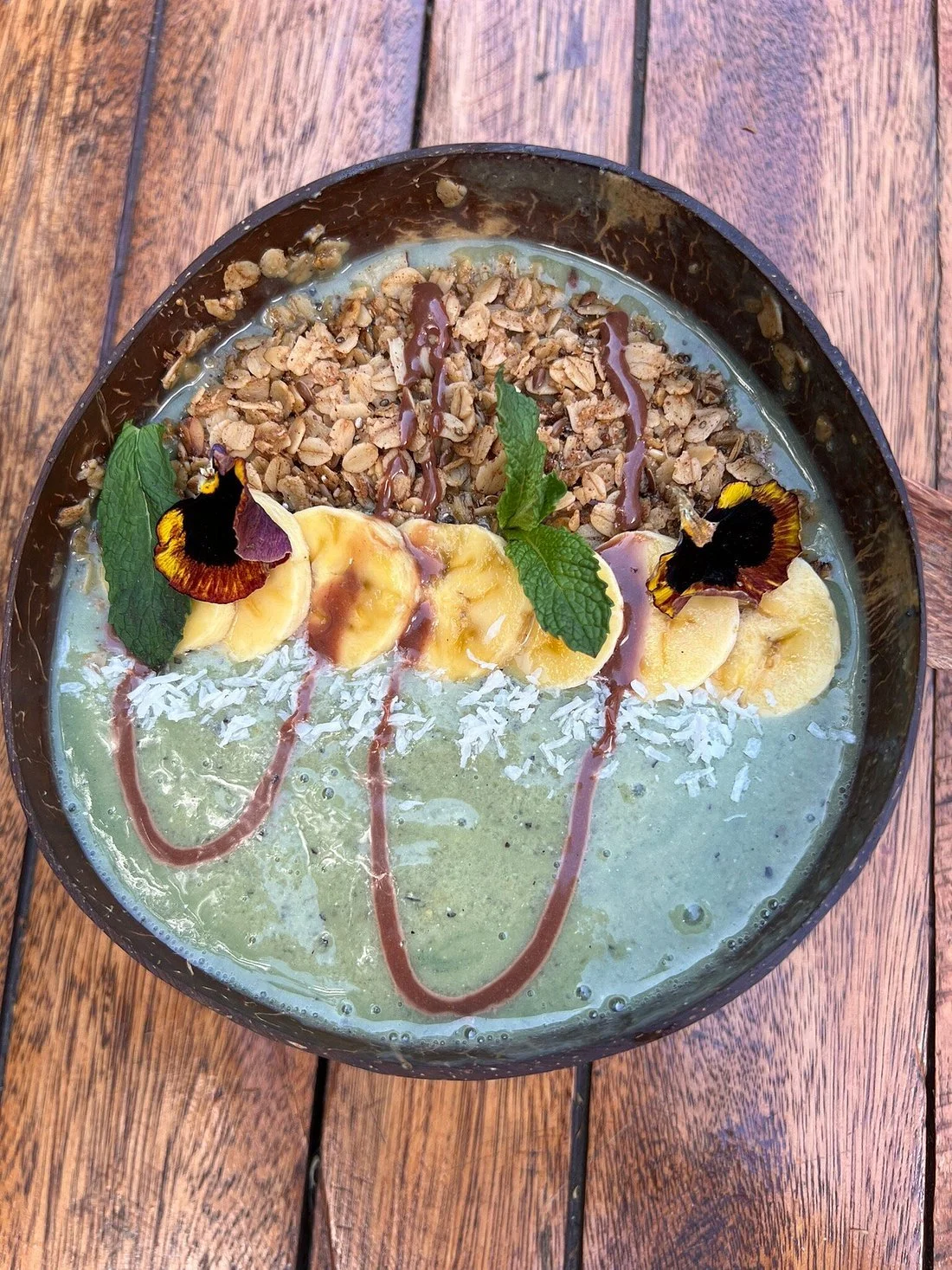 A green smoothie topped with sliced bananas, shredded coconut, granola, mint leaves, edible flower petals, and drizzled with chocolate and caramel sauces, served in a black bowl on a wooden surface.