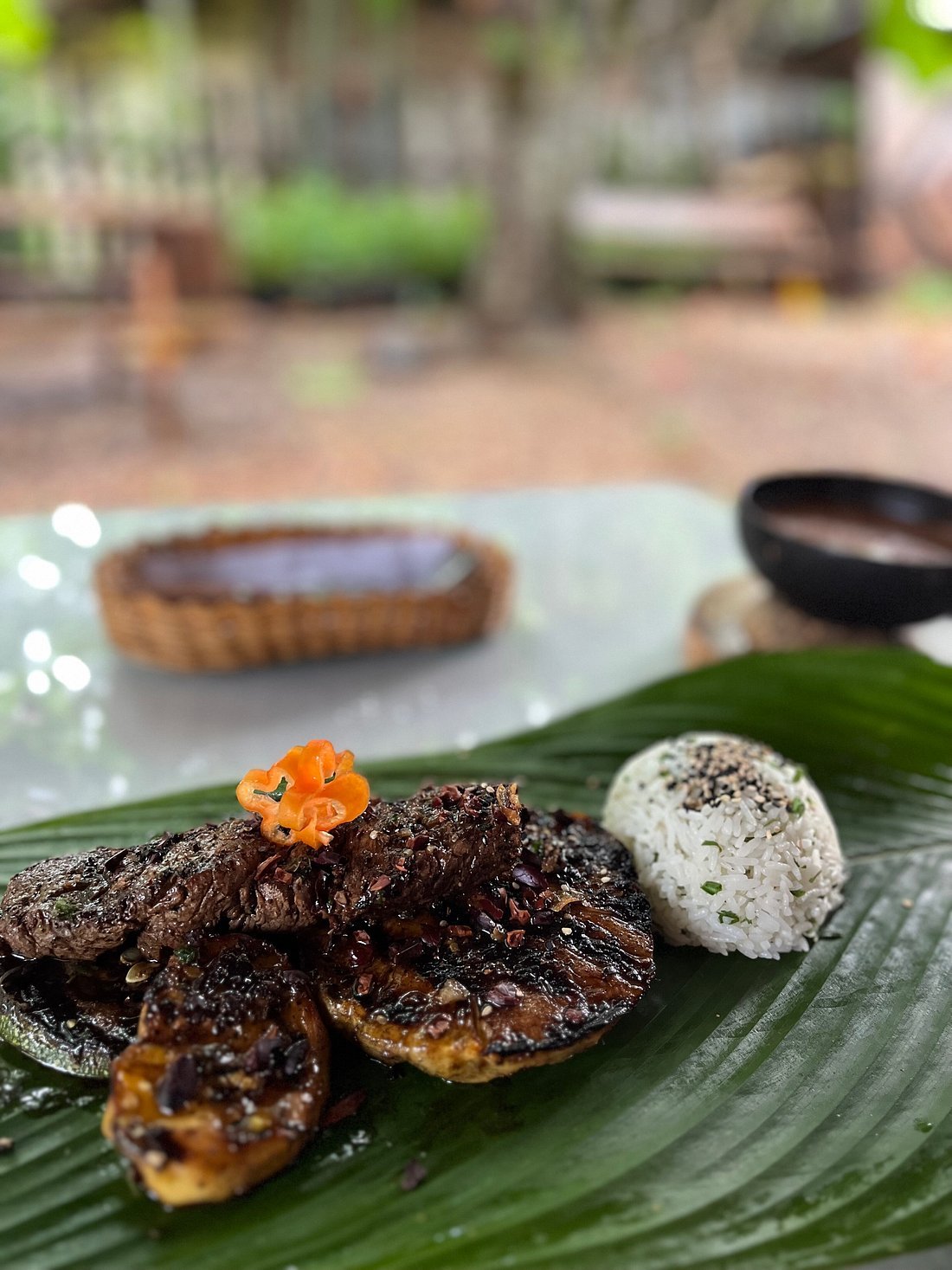 A plate with grilled meat and a scoop of rice on a banana leaf, garnished with a flower-shaped chili peel.