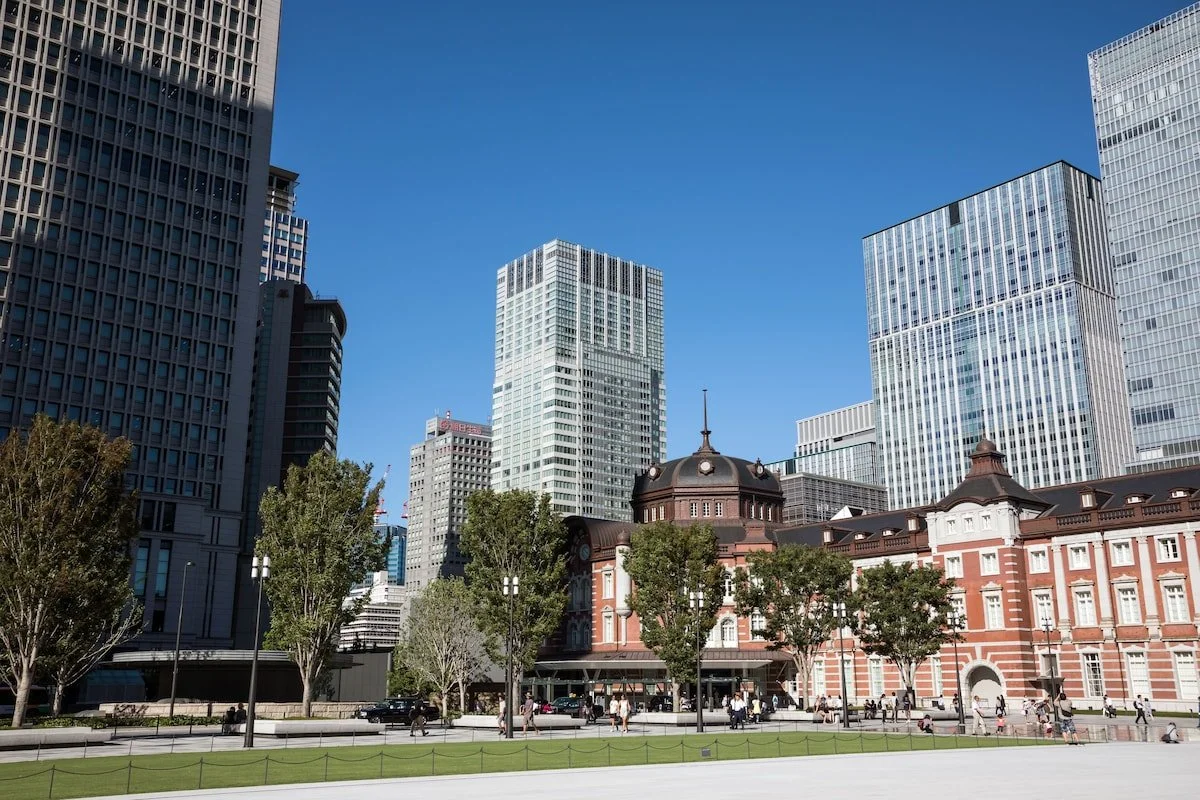 A cityscape featuring modern tall skyscrapers behind a historic red brick building with trees in front, on a clear blue sky day.