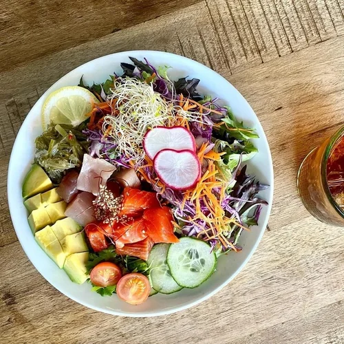 A colorful bowl of assorted fresh vegetables, fruits, and fish salad with lemon slices, radishes, cucumber, grape tomatoes, and sprouts on a wooden table, with a glass of iced tea beside it.