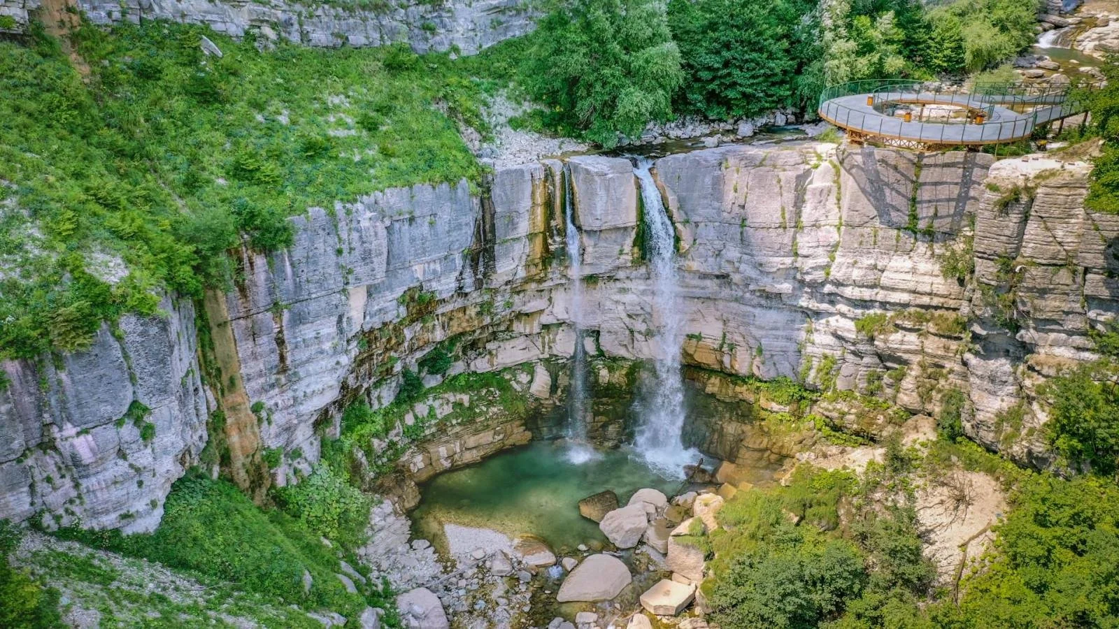 A scenic view of a waterfall inside a canyon with lush greenery surrounding it. There is a circular observation deck with a railing on the right side overlooking the waterfall.