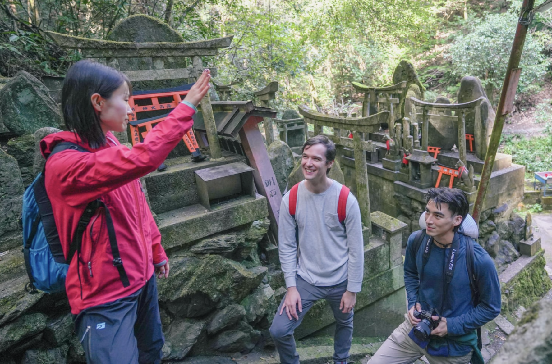 Three people, two men and one woman, standing in a forest near a Japanese shrine, with the woman giving a high-five to one of the men. The other man is holding a camera, and all are smiling.