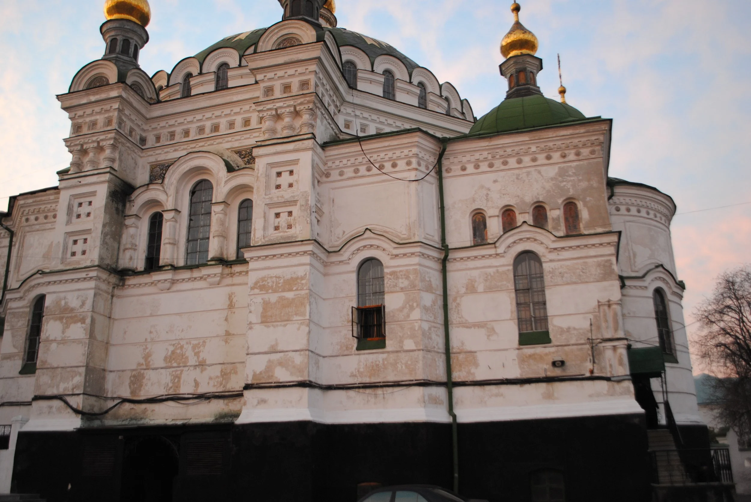 An old white church with green and gold domes, several arched windows, and decorative architectural details, set against a partly cloudy sky.