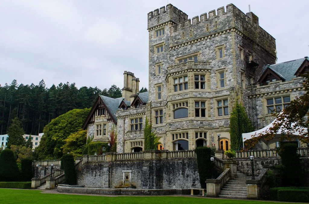 A large medieval-style stone castle with multiple towers and turrets, surrounded by a well-maintained lawn and trees, with an overcast sky overhead.