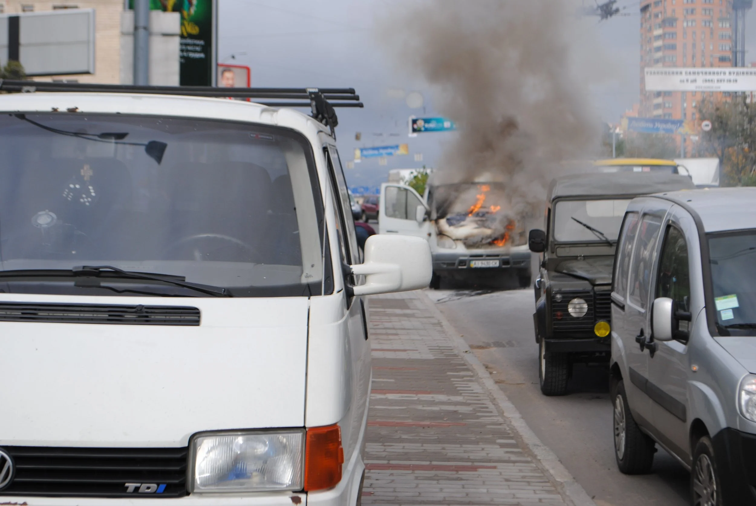 A street scene showing vehicles, including a white van and a gray car, with one vehicle in the background on fire and emitting thick black smoke. Kiev November 2011, Marina Parrish ©