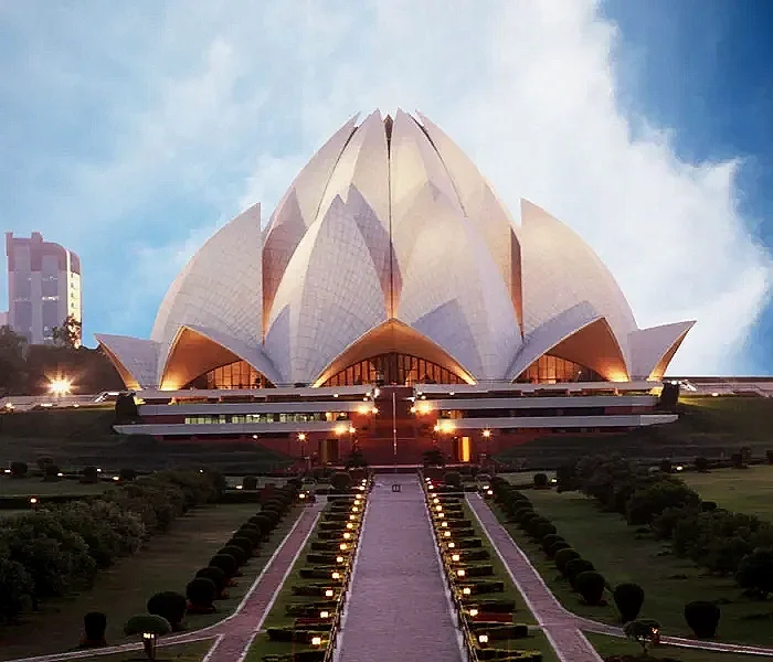 The Lotus Temple in Delhi, India, at dusk, with illuminated pathways leading to it and a partly cloudy sky background.