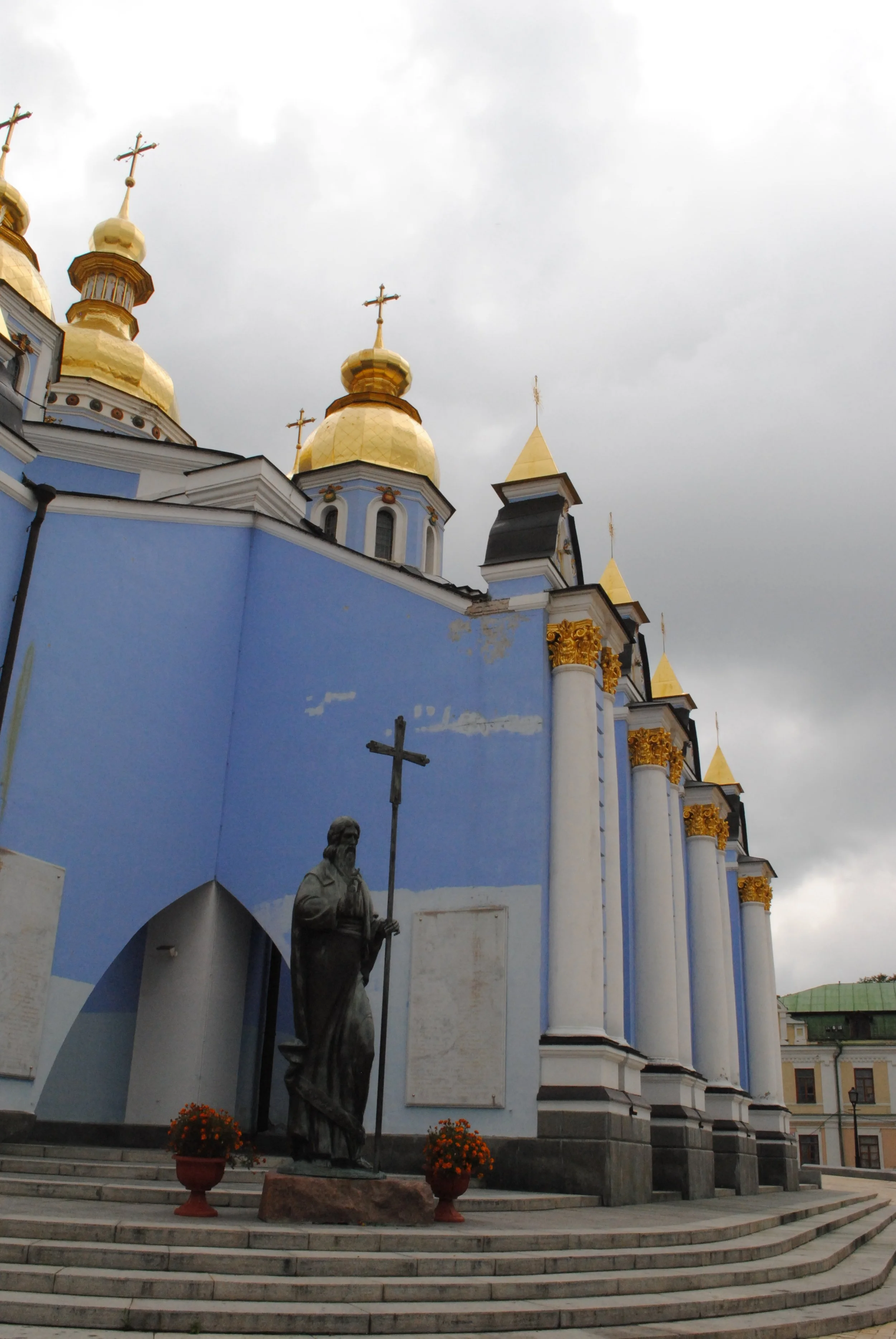A church with blue walls and gold domes topped with crosses, and a statue of a saint holding a staff, on a set of curved stairs.
