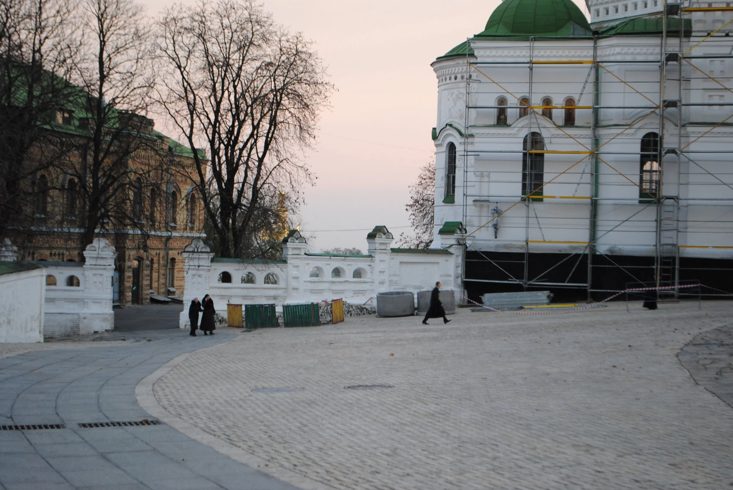 An outdoor scene with historic buildings, including one under renovation with scaffolding, leafless trees, a pathway with people walking, and a white wall with decorative arches.