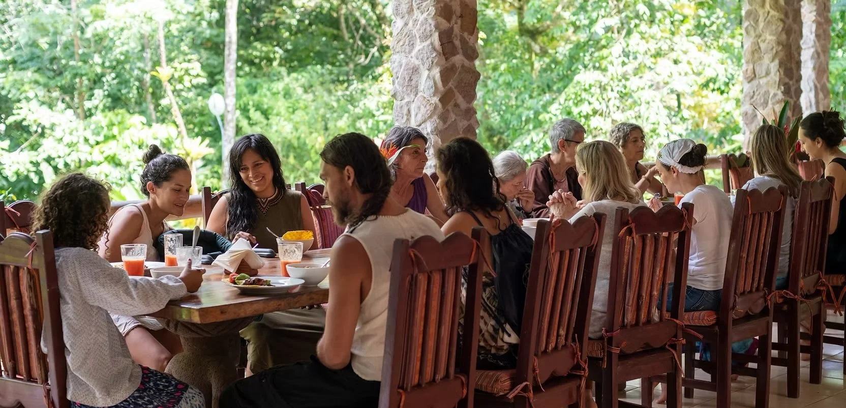 A diverse group of people, including children and adults, are sitting around a large dining table outdoors, enjoying a meal together amid green foliage and stone pillars.
