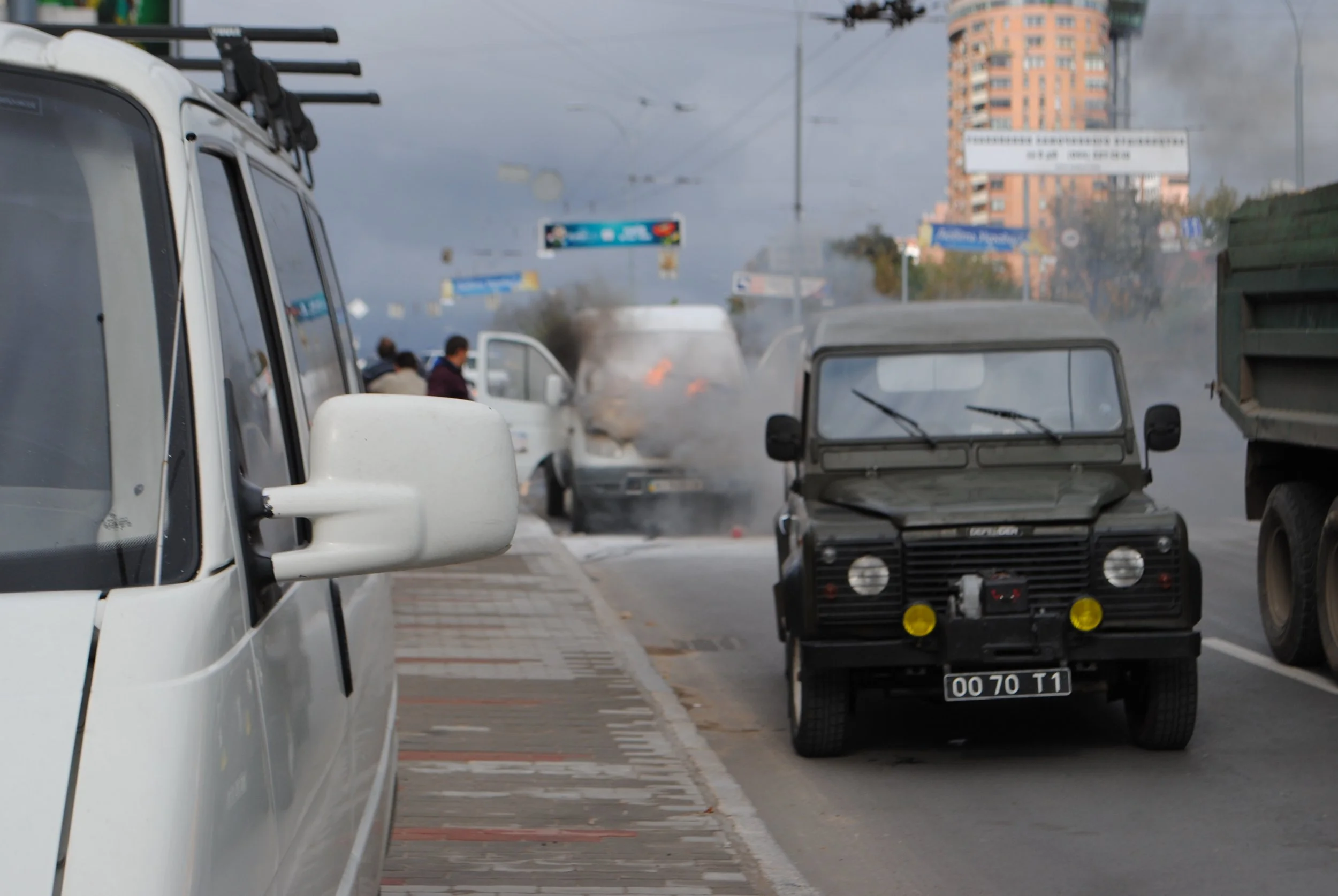 Several vehicles parked along a city street on a cloudy day, including a white van in the foreground, a black trailer, and other cars. There is smoke and fire emitting from a vehicle further down the street, and pedestrians are standing nearby. Build