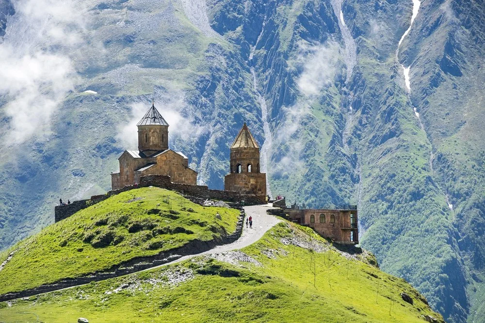 Ancient stone church with two towers on a grassy hilltop, surrounded by mountains.