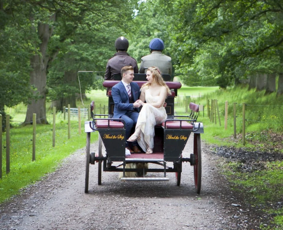 A couple in wedding attire riding in a horse-drawn carriage on a gravel path through a lush, green park or woodland area, with two men sitting in the back of the carriage.
