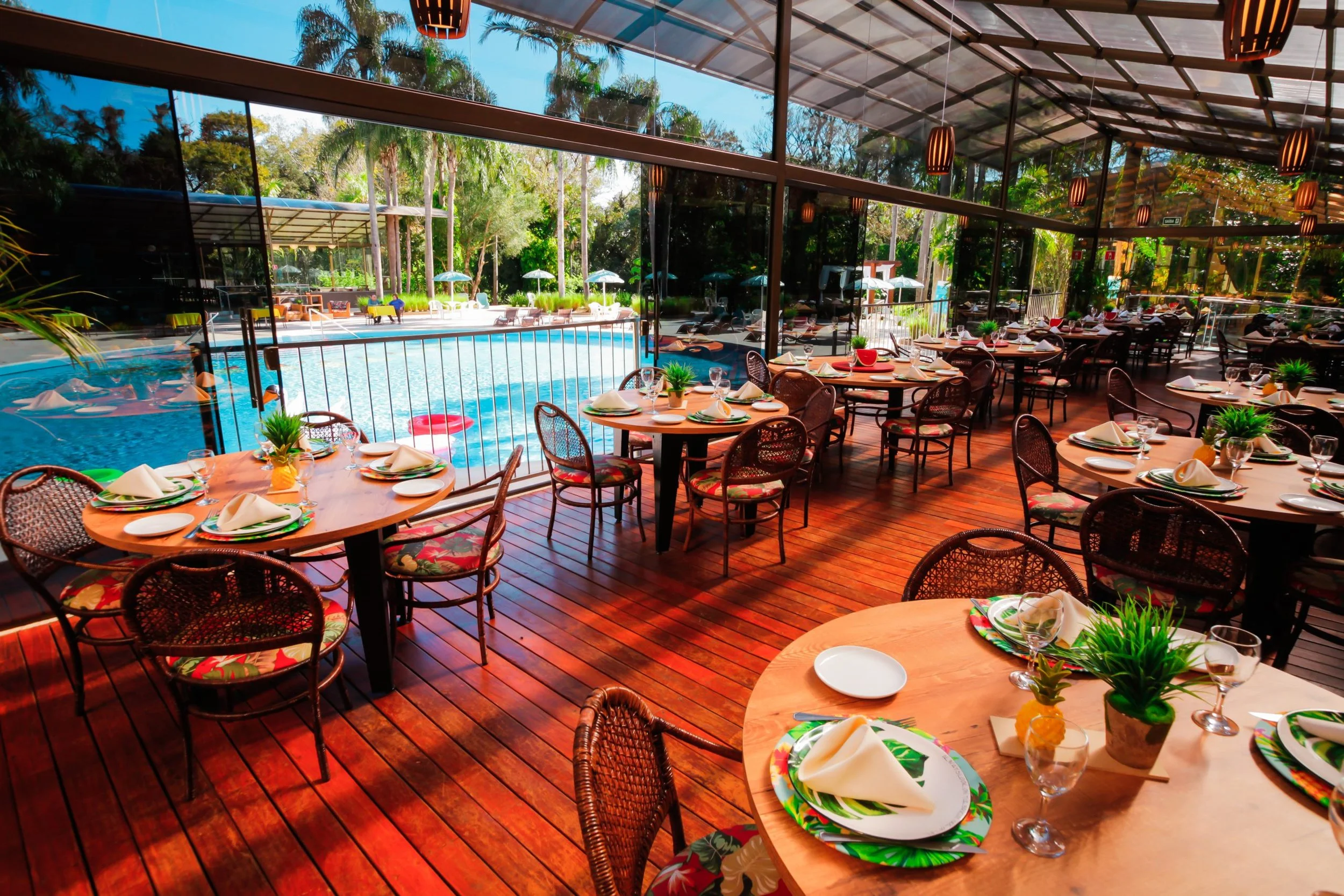 Indoor restaurant with round tables set with white napkins, glasses, and plates, overlooking an outdoor pool area with lounge chairs, umbrellas, and lush greenery.