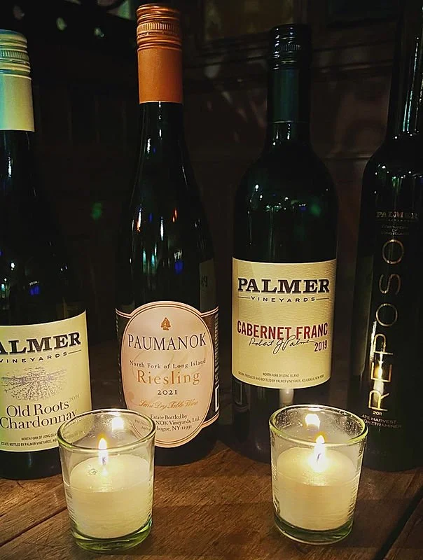 Four bottles of wine on a wooden surface, with two lit candles in front. From left to right: a Palmer Vineyards bottle of Old Roots Chardonnay, a Paumanok North Fork Long Island Riesling 2021, a Palmer Vineyards Cabernet Franc 2019, and a partially visible bottle of Opus One.