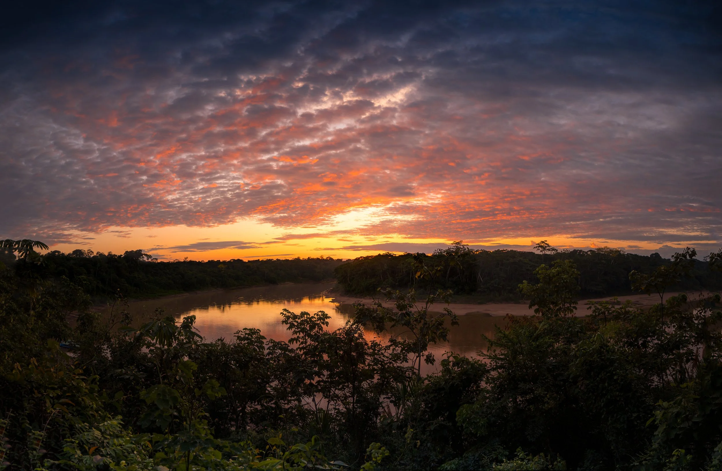 A sunset over a river with a cloudy sky and lush green trees in the foreground.