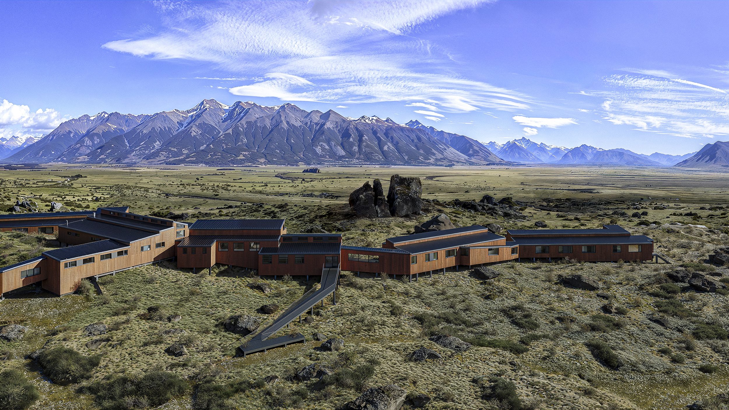 Modern wood-clad buildings on a rocky landscape with distant snow-capped mountains and a bright blue sky with wispy clouds.