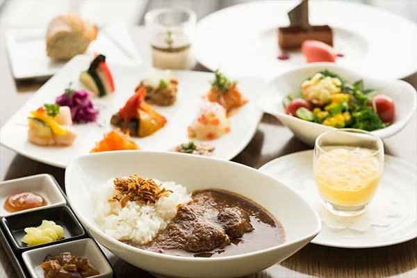 Japanese meal featuring rice with beef curry, assorted sashimi on a plate, salad with cherry tomatoes and lettuce, a glass of yellow beverage, and small dishes of condiments.