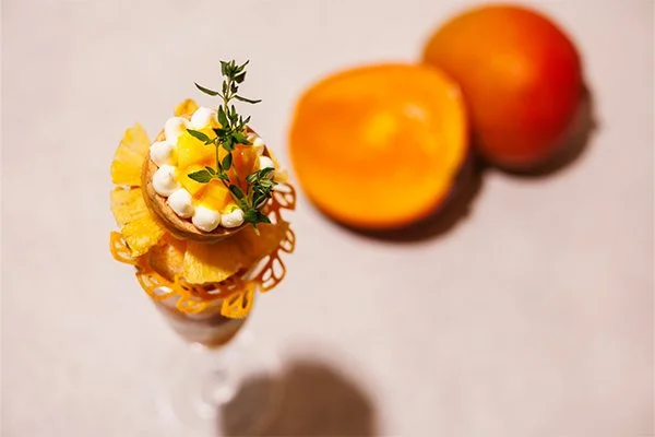 An appetizer in a tall glass with a fried chip, topped with a dollop of cream, yellow sauce, and garnished with herbs, next to a halved fresh peach and a whole peach.