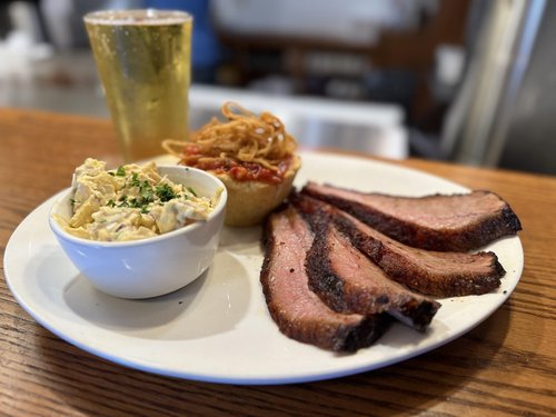 A plate of smoked meat with three slices, a cup of potato salad garnished with herbs, a small bread bowl of pulled pork with barbecue sauce, and a glass of beer in the background.