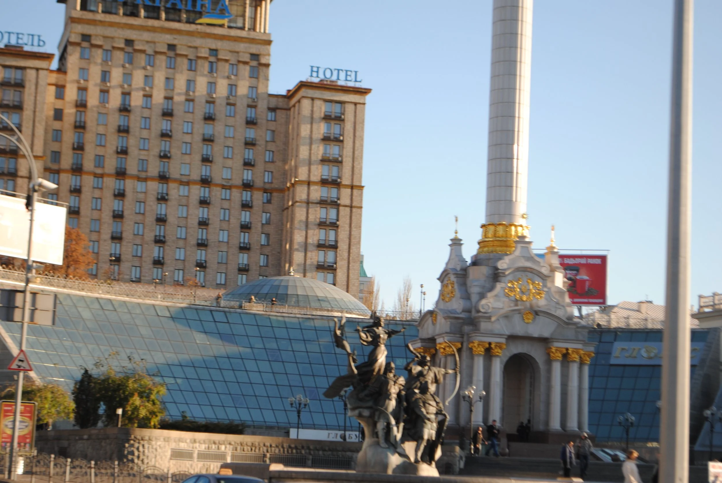 Tall building with a sign that says 'HOTEL' on top, a monument with a golden top and detailed statues, and a modern glass roof structure below. Clear blue sky in the background.