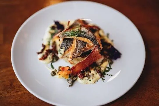 Plate of cooked fish with herbs, vegetables, and garnishes on a white dish.