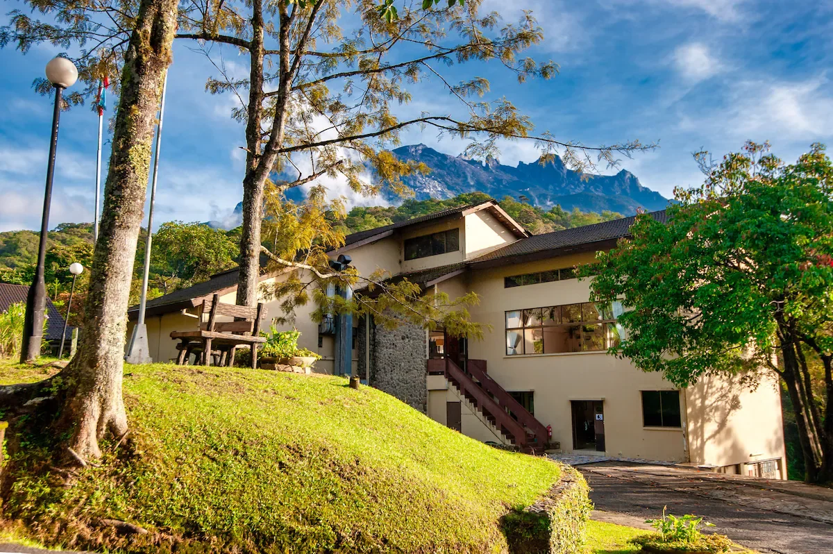 A house surrounded by trees on a grassy hill with mountain peaks in the background under a blue sky.
