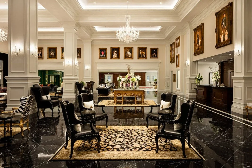 Elegant hotel lobby with black marble floors, white walls, ornate crown molding, a large chandelier, and framed classical paintings, featuring black armchairs, a gold table with floral arrangements, and a reception desk.