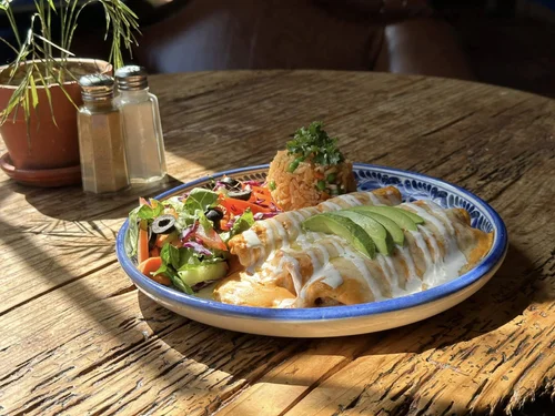 Plate of Mexican food with enchiladas topped with avocado slices, side salad with olives and vegetables, and a scoop of Mexican rice, on a rustic wooden table, with sunlight and a small potted plant in the background.