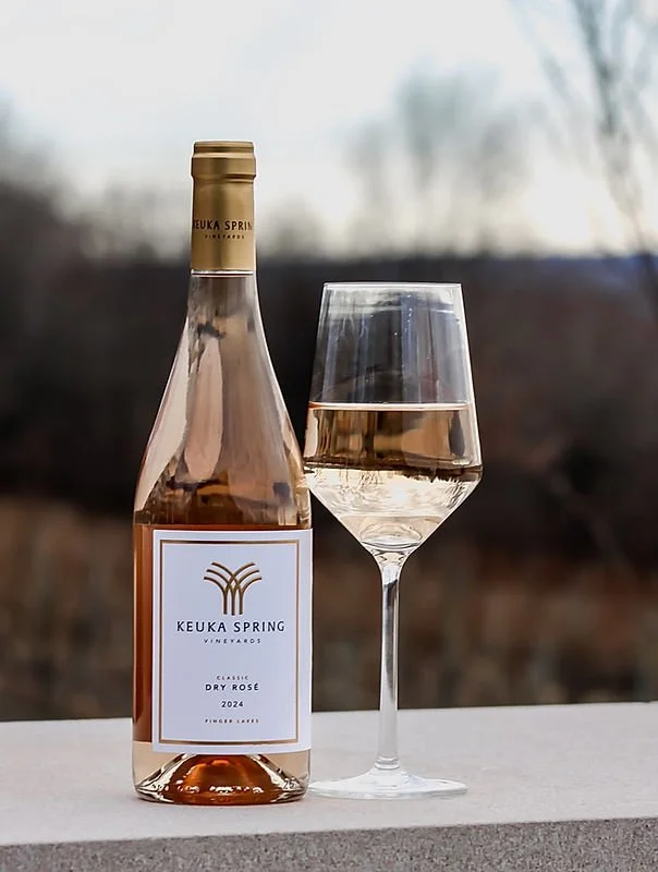 A bottle of Keuka Spring Vineyards dry rosé wine next to a filled wine glass on a white surface, with an outdoor background of trees and sky.