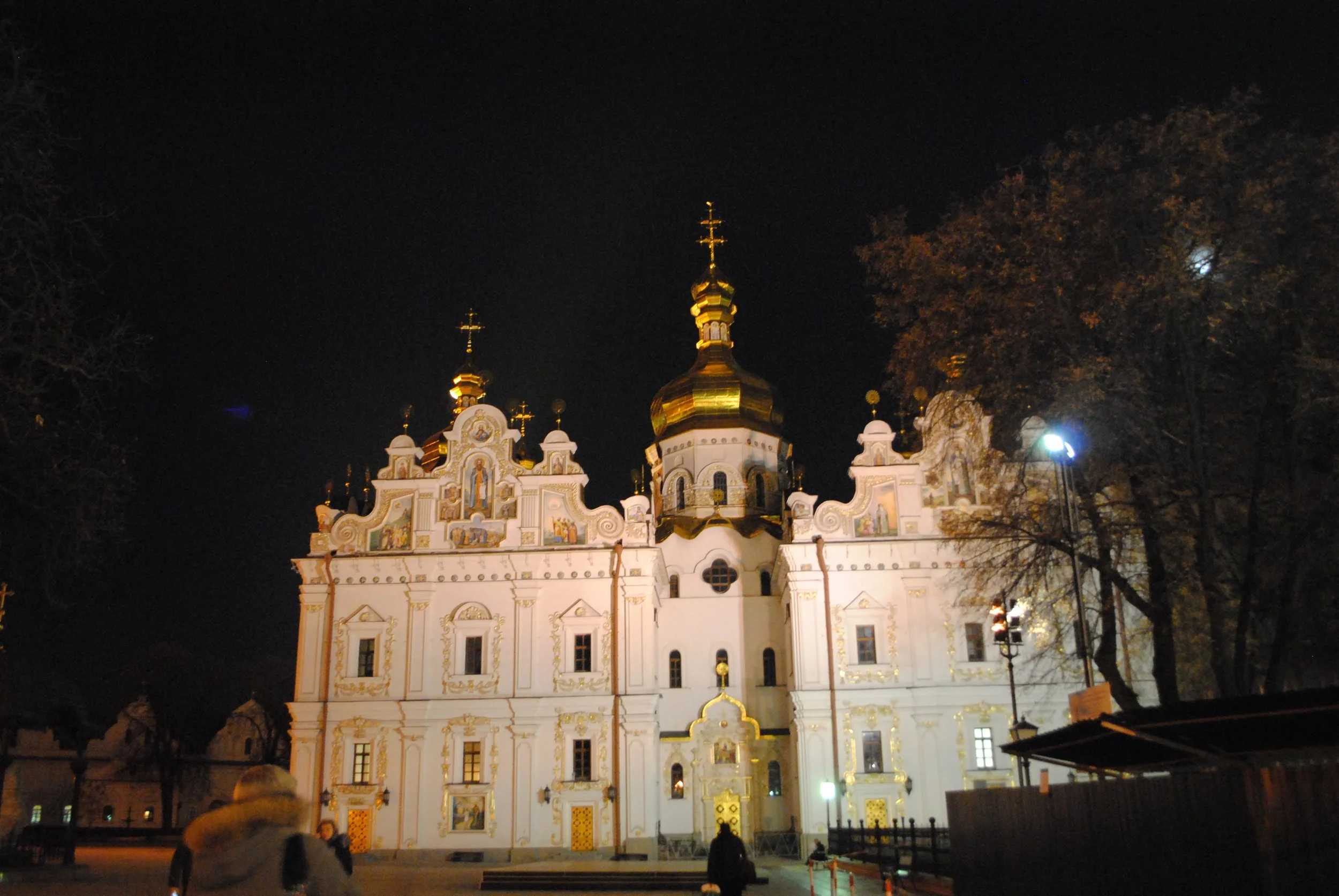 A historic church building at night with ornate white and gold architecture, illuminated lighting highlights its domed golden roofs and intricate details. There are trees and a dark night sky in the background.