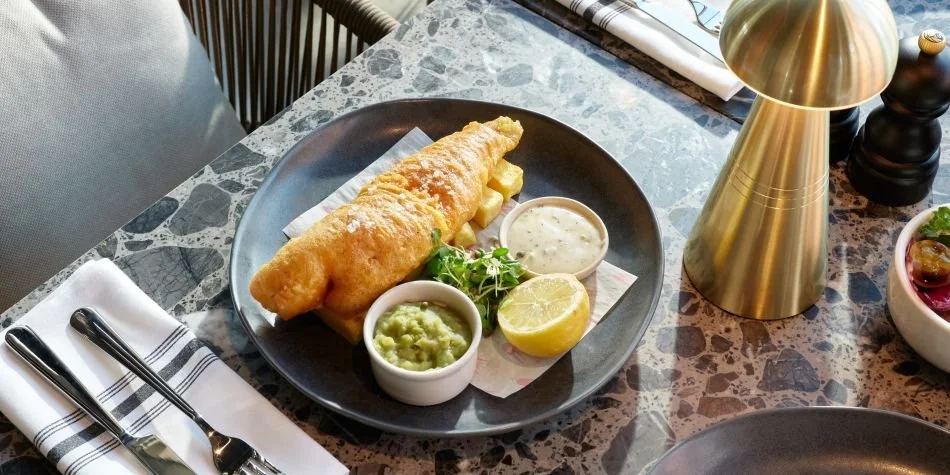 Fried fish fillet with tartar sauce, guacamole, and lemon on a black plate at a restaurant table.