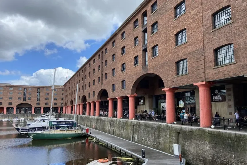 View of a brick building with multiple windows and arched passageways, with red columns supporting the upper levels. In front, there is a dock with boats and people walking along the waterfront.