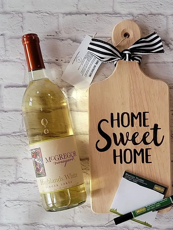 A bottle of McGregor Vineyards Highlands White wine next to a wooden cutting board with the words "Home Sweet Home" written on it, a business card, and a green and black pen, all arranged against a white brick wall background.